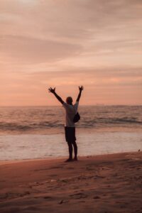 man in white shirt and black shorts standing on seashore during sunset